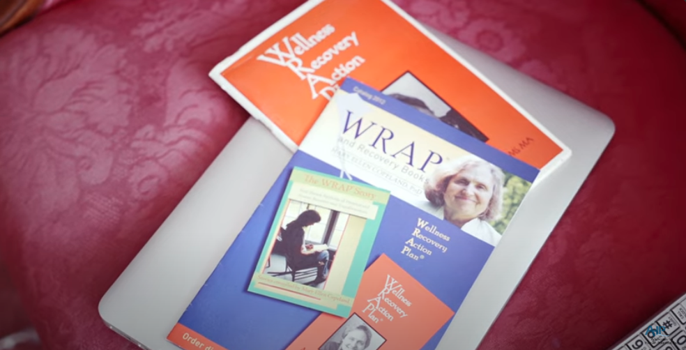 A set of Wellness Recovery Action Plan (WRAP) booklets are spread out on a cushioned chair. One booklet features a photo of a smiling person on the cover. A laptop is partially visible underneath the booklets. This is the thumbnail image for a video.